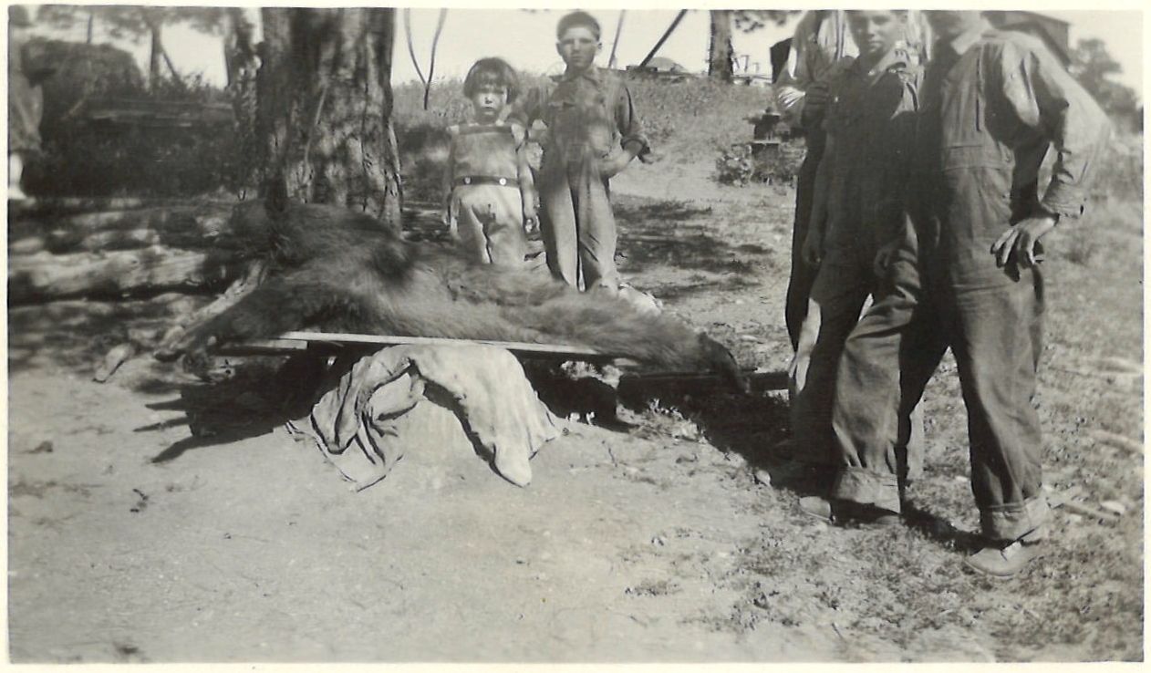 Children and ranch hands gathered around a bear laid on a plank bench
