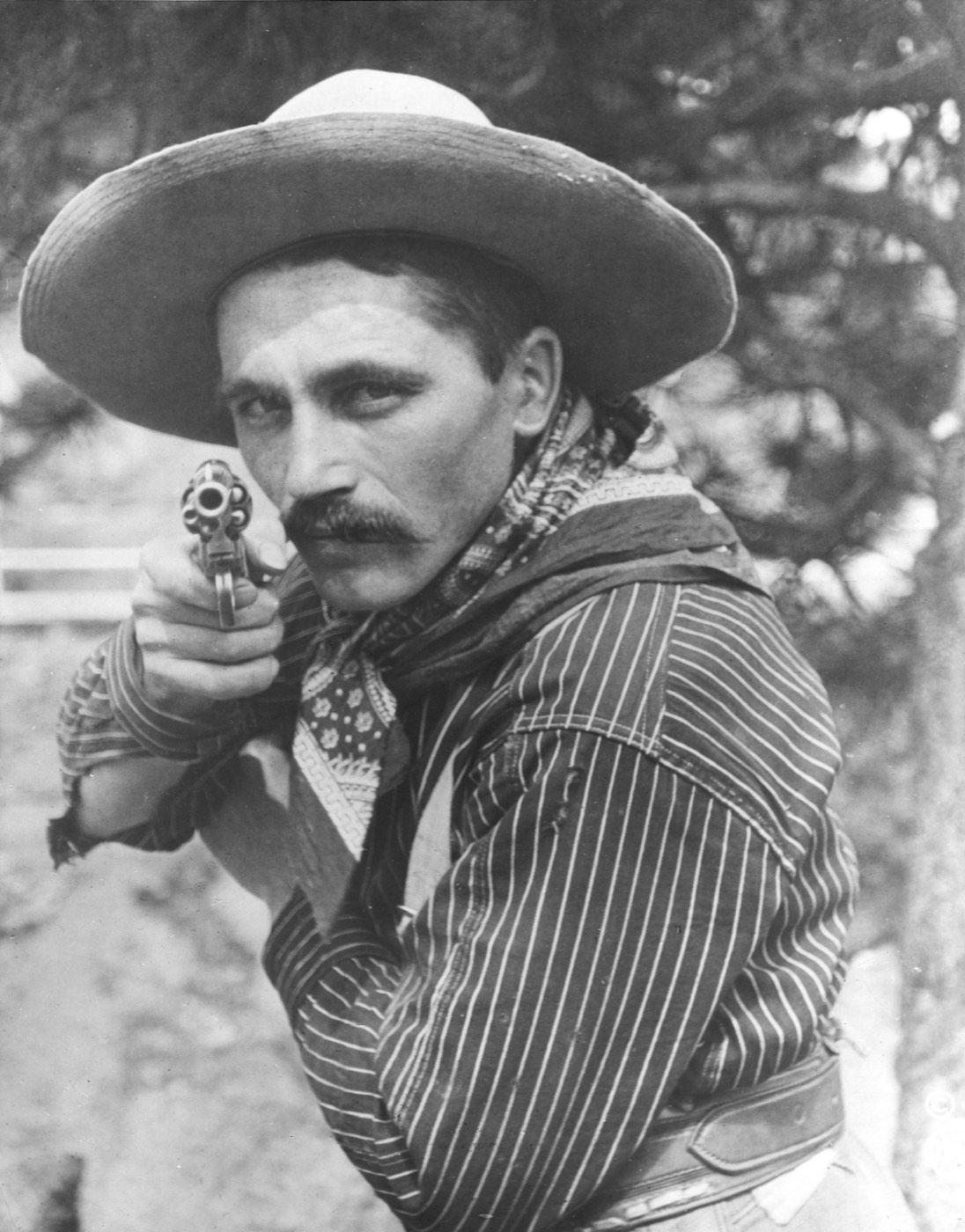 Undated portrait of Steve Pastore in a wide-brimmed hat and neckerchief, sighting a revolver directly at the camera