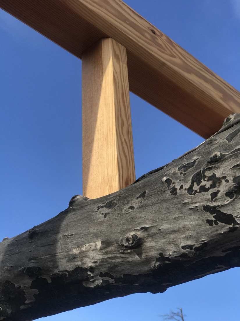 Timber post-and-beam joint against a weathered fire-scarred log rail and blue sky